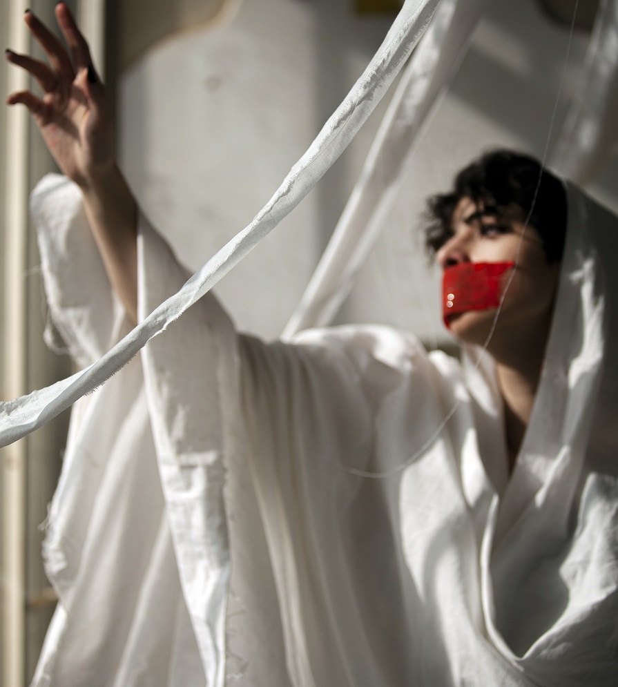 femme voilée de blanc, un masque en tissu rouge sur la bouche levant la main vers une fenêtre