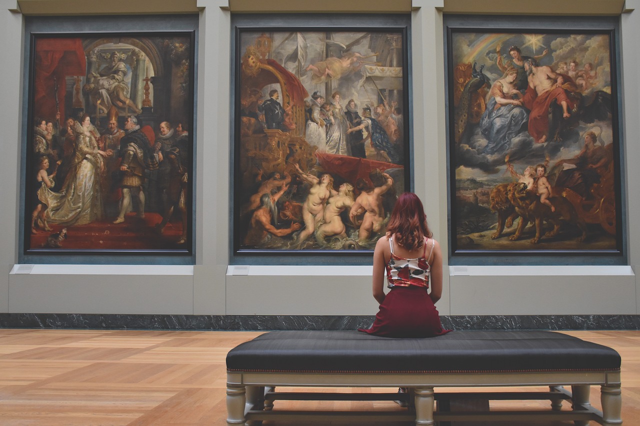 femme vue de dos, assise devant des tableaux dans un musée