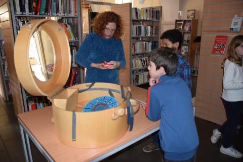 enfants face à une femme qui leur montre un objet rouge à côté de la malle ronde ouverte