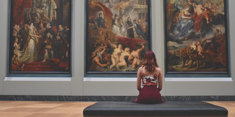 femme vue de dos, assise devant des tableaux dans un musée